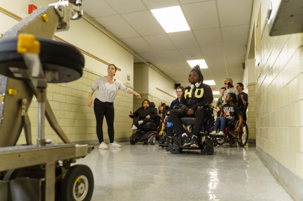 A group of students using wheelchairs look forward into a camera while a teacher is pointing toward the ground.