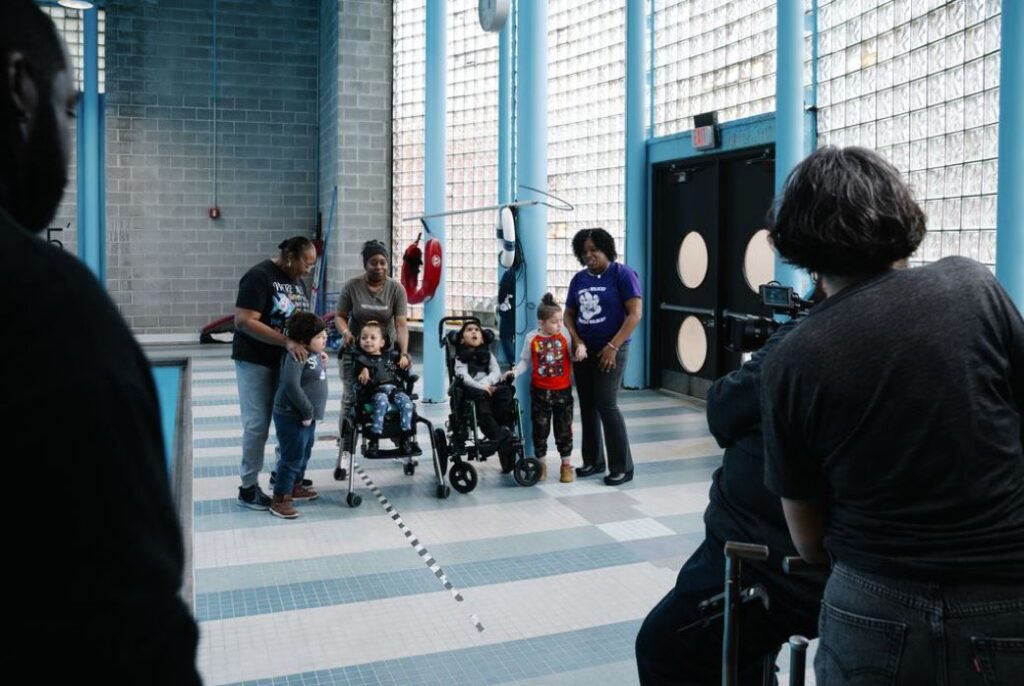 A group of young students, two using wheelchairs, stand with their teachers in a large open room with windows and a swimming pool. They are facing a camera and film crew.