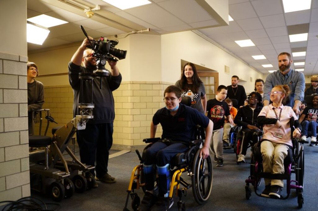 A group of students using wheelchairs and their teachers are moving forward passing by a camera and videographer with smiles on their faces.